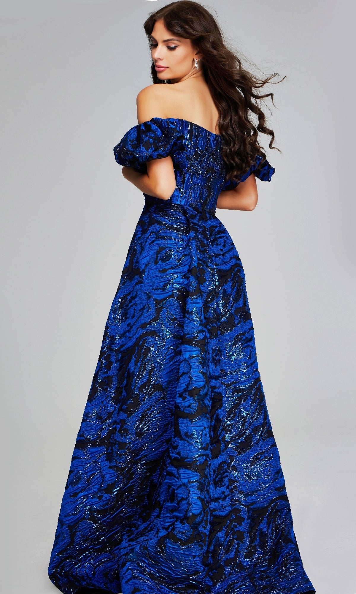 A woman stands facing away in a Jovani 40315 long formal dress, an off-the-shoulder, floor-length gown with blue and black patterns. Her long wavy hair cascades down her back.